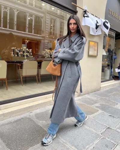 best long coats are shown in a photo of a long coat outfit with a woman standing on the sidewalk wearing a long gray peacoat over straight-leg jeans with chunky sneakers and a tan suede handbag