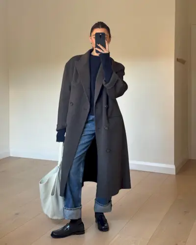 best long coats are shown in a photo of a long coat outfit with a woman standing in her home wearing a long gray coat over a navy sweater with baggy jeans, black loafers, and a white tote bag