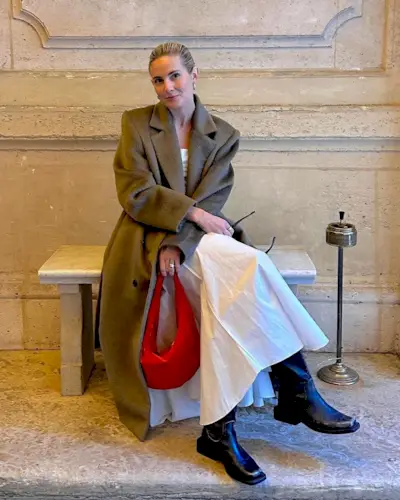 best long coats are shown in a photo of a long coat outfit with a woman sitting in a museum wearing a long tan coat over a white maxi dress with black knee-high riding boots and a red handbag