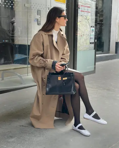 best long coats are shown in a photo of a long coat outfit with a woman sitting on a bus bench wearing a long trench coat over a white turtleneck with a mini skirt, black tights, white ballet flats, and a black handbag