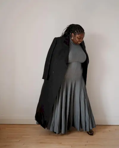 best long coats are shown in a photo of a long coat outfit with a woman standing inside her home wearing a long black coat over a gray maxi dress with black pointed heels