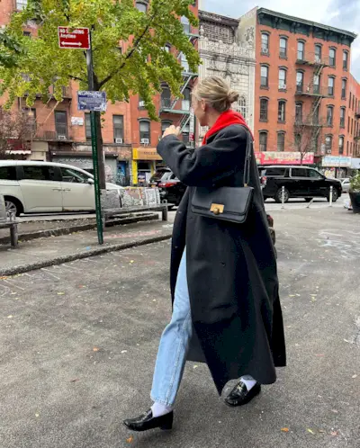 best long coats are shown in a photo of a long coat outfit with a woman standing outside wearing a red hoodie under a long black coat with straight-leg jeans, black loafers, and a black crossbody bag