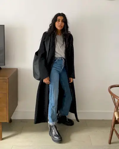 best long coats are shown in a photo of a long coat outfit with a woman standing in her home wearing a long black coat over a gray t-shirt with straight-leg jeans, black chelsea boots, and a black sling bag