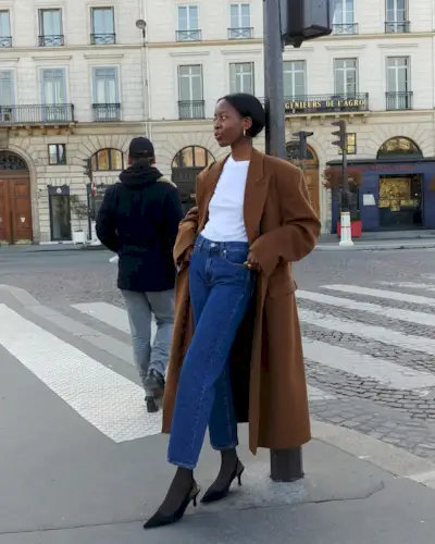 best long coats are shown in a photo of a long coat outfit with a woman standing outside wearing a long brown coat over a white t-shirt with straight-leg jeans, black tights, and black slingback heels