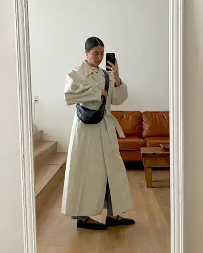 best long coats are shown in a photo of a long coat outfit with a woman standing in her home wearing a long tan trench coat, a sling bag, and black mary jane flats