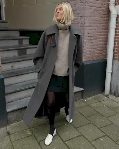 best long coats are shown in a photo of a long coat outfit with a woman standing outside wearing a long gray coat over a tan turtleneck with a black mini skirt, black tights, and white loafers