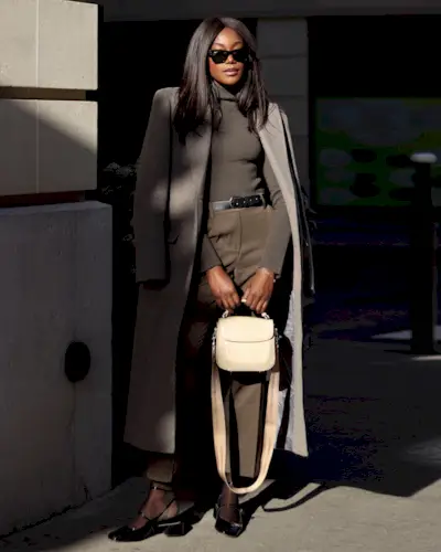 best long coats are shown in a photo of a long coat outfit with a woman standing outside on a sidewalk wearing a long gray coat over a dark gray turtleneck sweater with tan trousers, black Mary Jane flats, and a white handbag