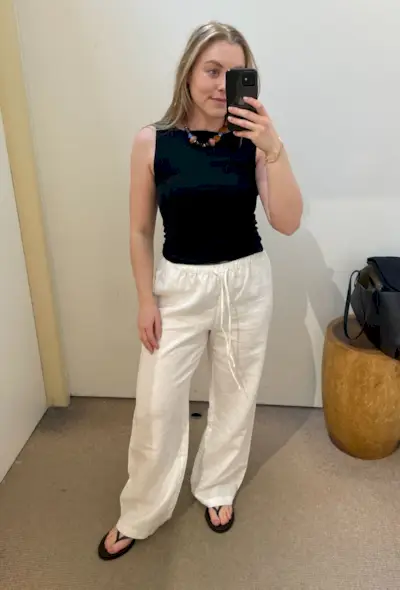 Woman wears black top, bead necklace, white linen trousers and black sandals
