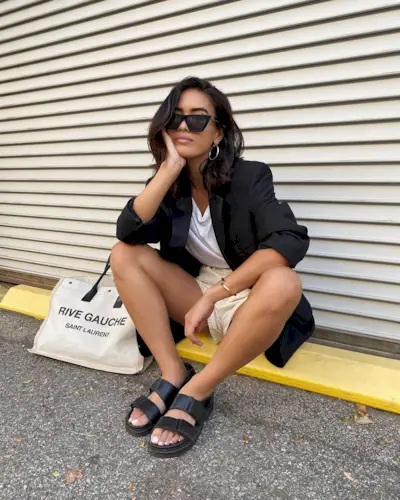 A woman wearing shorts, a white shirt, a black blazer, black sandals, large sunglasses, and a Saint Laurent tote.
