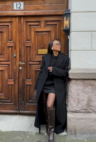 A woman standing outside in a doorway wearing a long black coat over a black crewneck sweater, black miniskirt, brown knee-high boots, brown handbag, and gold earrings.