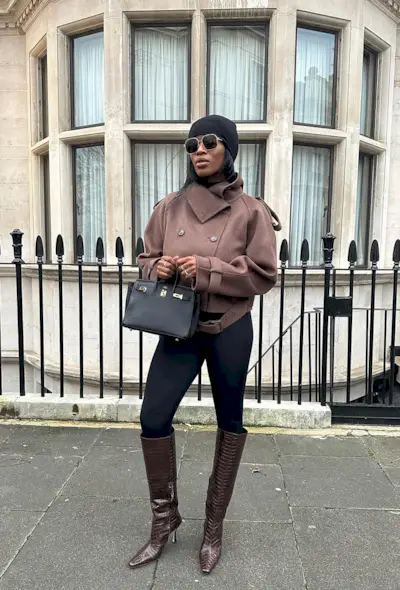 A woman standing on the sidewalk wearing a black beanie, sunglasses, a brown cropped coat, brown sweater, black leggings, brown knee-high boots, and a black handbag.