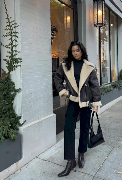 A woman wearing a black turtleneck, a brown leather shearling jacket, black jeans, brown ankle boots, and a black bag.
