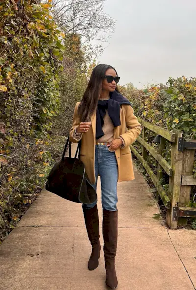 A woman standing in a garden wearing a tan blazer, a tan sweater, skinny jeans, brown suede knee boots, and a brown handbag.