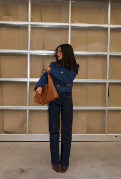 A woman standing outside wearing a jean jacket with dark wash straight-leg jeans, brown ankle boots, and a brown slouchy bag.