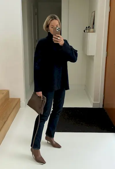 A woman taking a mirror selfie wearing a navy blue turtleneck, dark wash jeans, brown ankle boots, and a brown clutch bag.