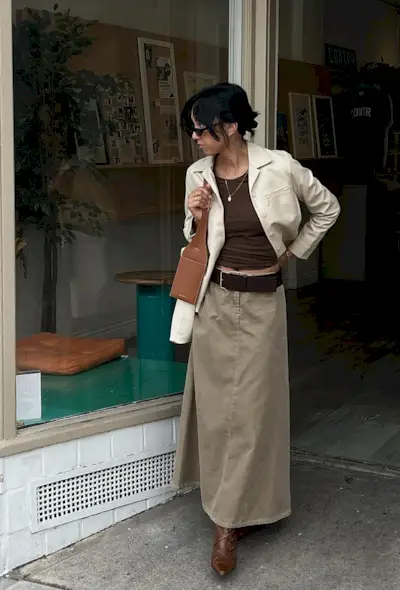 A woman standing outside a coffee shop wearing a tan barn jacket, a chocolate brown t-shirt, a long tan cargo skirt, brown handbag, and brown knee-high boots.