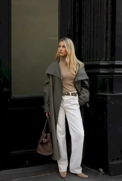 A woman standing on the sidewalk wearing an olive green trench coat, tan turtleneck, white jeans, studded brown belt, suede ankle boots, and an oversize handbag.
