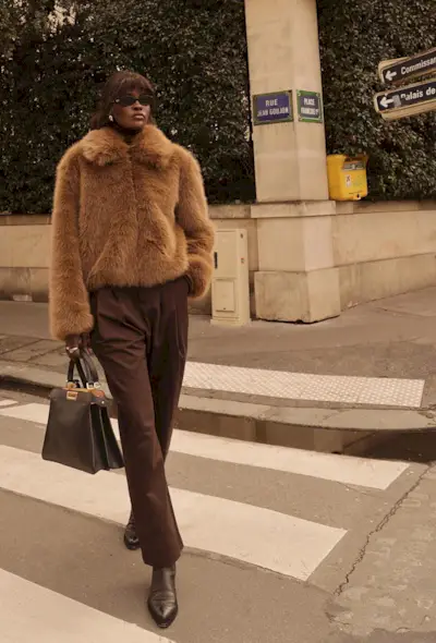 A woman standing on the sidewalk wearing a light brown cropped fur coat with dark brown trousers, brown ankle boots, and a brown handbag.