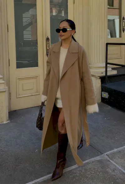 A woman walking on the sidewalk wearing a long tan coat over a white mohair crewneck, a white miniskirt, chocolate brown knee-high boots, and a brown handbag.