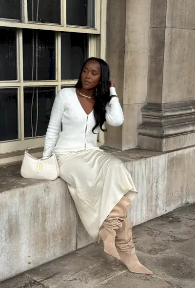 A woman sitting on a windowsill wearing a white cardigan, white slip skirt, tan slouchy suede boots, and a white shoulder bag.