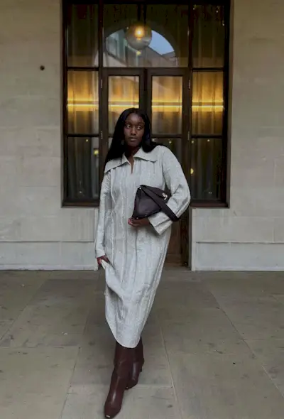 A woman standing outside wearing a gray sweaterdress, brown knee-high boots, and a brown handbag.
