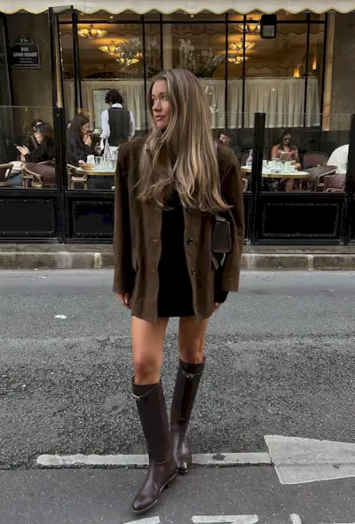 A woman standing in the street wearing a brown suede jacket over a black minidress, brown handbag, and brown knee-high boots.