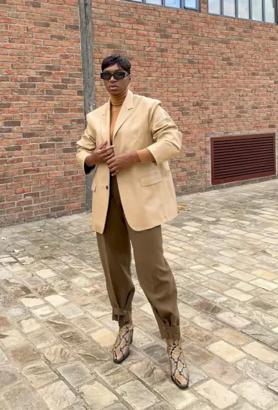 A woman standing outside wearing a light tan blazer over a turtleneck, olive green trousers, and snakeskin ankle boots.