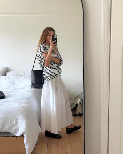 British fashion influencer taking a mirror selfie with a striped T-shirt, white maxi skirt, woven bucket bag, and loafers