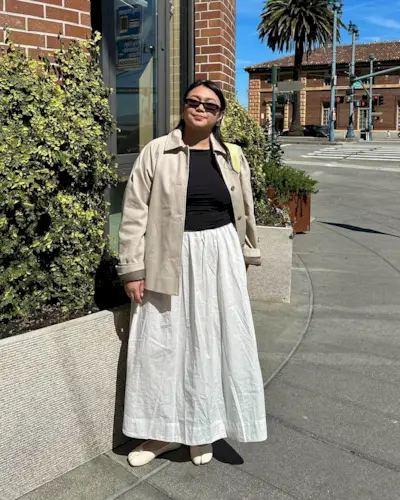 Californian influencer Marina Torres poses on a sunny street in a neutral jacket, black top, white poplin skirt, and Margiela ballet flats