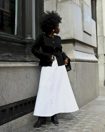 fashion influencer Danielle Oreoluwa Jinadu poses on the street wearing a black sweater-jacket, full white maxi skirt, and ankle boots