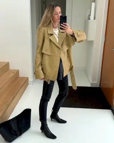 influencer Anouk Yve poses for a mirror selfie wearing a cropped trench coat, white top, skinny black jeans, and black square-toe kitten heel boots