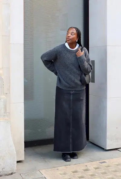 A denim skirt outfit is shown in a photo of a woman on the sidewalk wearing a white t-shirt under a gray sweater styled with a gray denim maxi skirt, black boots, and a black shoulder bag