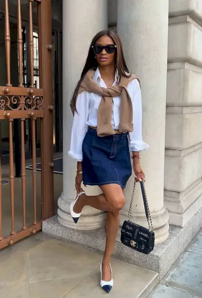 A denim skirt outfit is shown in a photo of a woman on the street wearing a white button-down shirt with a brown tied cardigan over the shoulders styled with a denim mini skirt, brown belt, chanel bag, and two-tone slingback heels