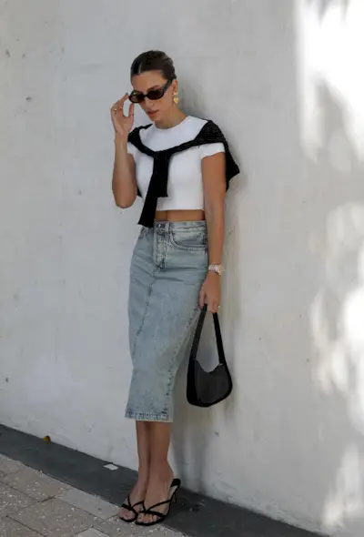A denim skirt outfit is shown in a photo of a woman leaning against a wall wearing gold earrings, black sunglasses, a cropped white t-shirt, a cardigan tied around the shoulders, a distressed midi skirt, a black shoulder bag, and black mules