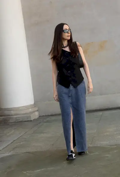 A denim skirt outfit is shown in a photo of a woman on the street wearing tinted lenses, a beaded necklace, a black waistcoat, a denim maxi skirt, and black sneakers