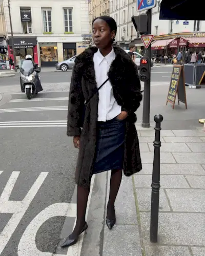 Sylvie wears a brown fur coat, white button-down, denim skirt, sheer tights, and slingbacks.