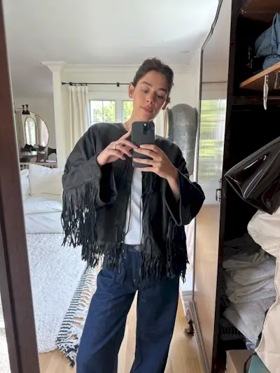 Ilana wears a black leather fringe jacket, white tee, and blue jeans.
