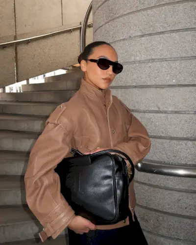 Ingrid wears a tan leather bomber jacket with sunglasses and a black bag.