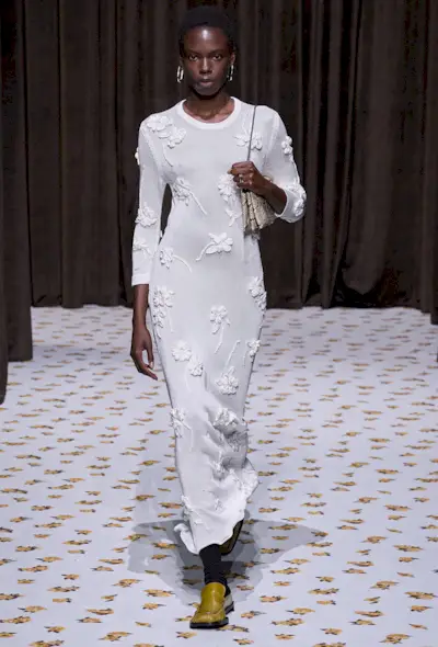A snakeskin purse is shown in a photo of a model walking down Jil Sander's spring runway shows carrying a python shoulder bag styled with silver earrings, a white knit dress, and yellow loafers