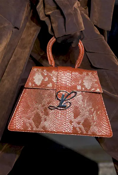A snakeskin purse is shown in close-up photo of a model walking down the Luar spring 2025 runway show carrying a brown python handbag with a brown trench coat