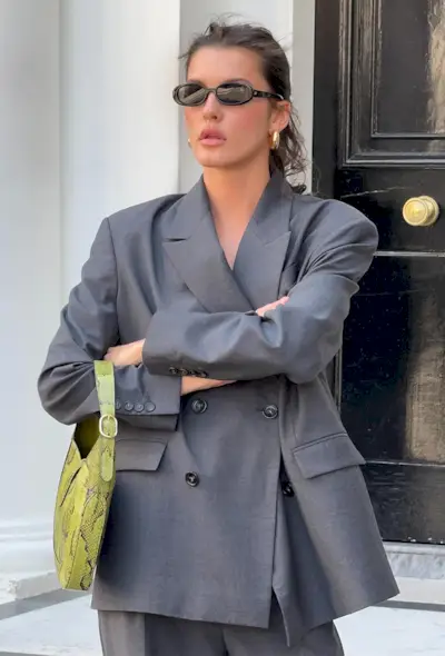 A snakeskin purse is shown in a street-style photo of a woman standing on a stoop wearing black sunglasses, gold hoops, a gray blazer with matching trousers and a green snakeskin shoulder bag
