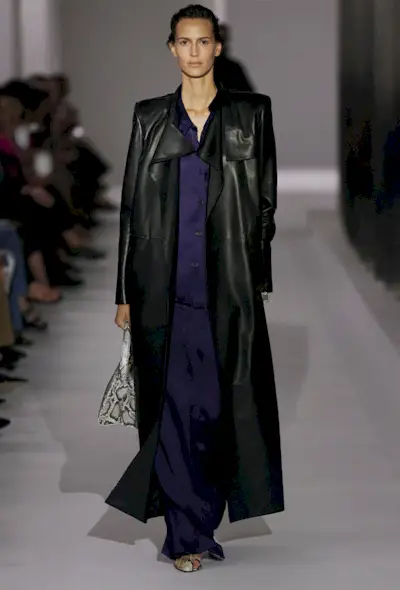 A snakeskin purse is shown in a photo of a model walking down the of Khaite's spring 2025 runway show carrying a python handbag with a a long black leather trench coat, navy blue satin shirt, navy satin trousers, and snakeskin heels