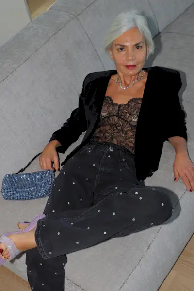 a casual party outfit shown on a woman laying on a couch wearing a black velvet blazer over a lace bodysuit with black jeans and sparkly shoes and a rhinestone bag