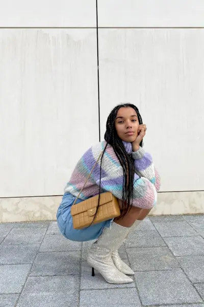 a casual party outfit shown on a woman standing outside on a sidewalk wearing a striped sweater with denim long shorts and glitter knee-high boots and a chanel shoulder bag