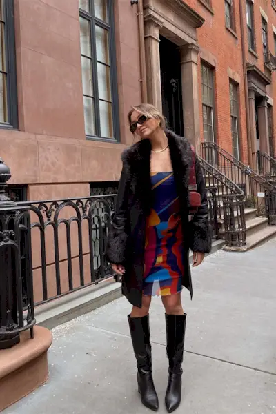 a casual party outfit shown on a woman standing outside a brownstone wearing a black trench coat with fur trench coat layered over a strapless tube print dress with black knee-high boots
