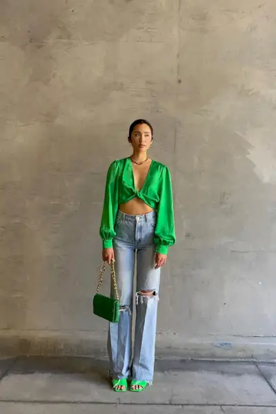 a casual party outfit shown on a woman standing outside wearing a green satin cropped blouse with baggy jeans, green mules, and a green shoulder bag