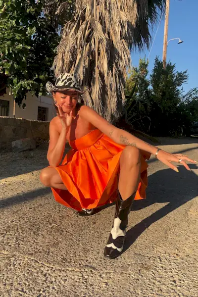 a casual party outfit shown on a woman standing outside wearing a zebra print cowboy hat with an orange mini dress and cowboy boots