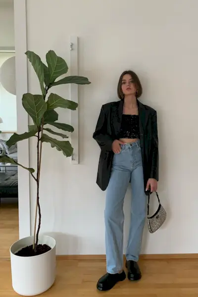 a casual party outfit shown on a woman standing inside their house wearing an oversize black blazer layered over a sequin crop top with baggy jeans and black ankle boots