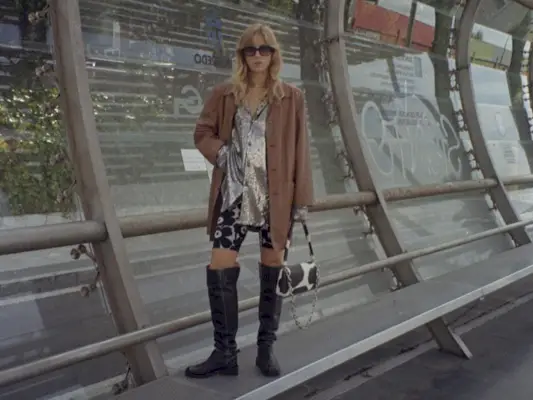 a casual party outfit shown on a woman standing at a bus stop wearing a tan blazer over a sequin button-down shirt with printed capri pants and over-the-knee moto boots