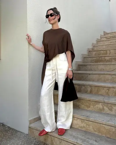 style influencer Annabel Rosendahl poses on an outdoor staircase wearing a brown asymmetrical cape top, white linen pants, a black bag, and red The Row jelly flats